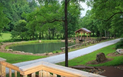 A serene pond surrounded by trees, with a wooden gazebo and a gravel path leading through a lush green landscape.