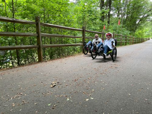 Two people riding recumbent tricycles on a tree-lined path, surrounded by greenery and a wooden fence.