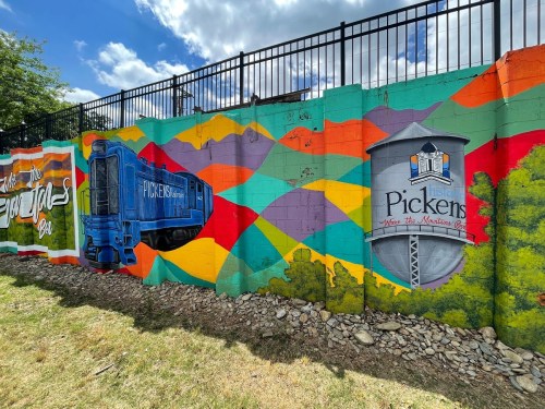 Colorful mural featuring a blue train and a water tower, set against a vibrant geometric background in Pickens.