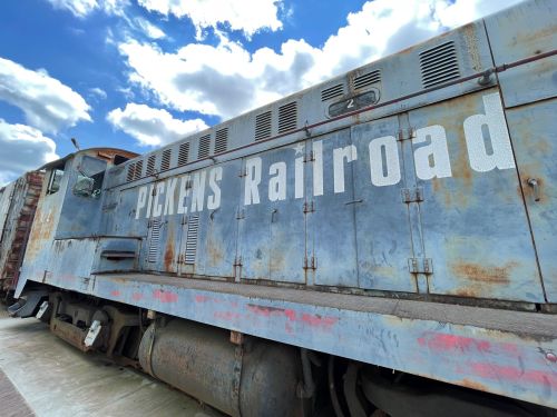 Close-up of a weathered train engine with "PICKENS Railroad" painted on the side, set against a blue sky with clouds.