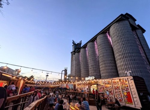 A lively outdoor market with string lights, food stalls, and large silos in the background during sunset.