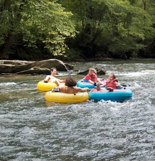 Four children float on colorful inner tubes down a river, surrounded by lush greenery and rocky banks.