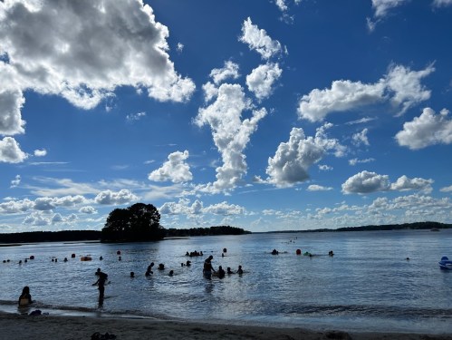 A sunny beach scene with people swimming in calm water, surrounded by fluffy clouds and a distant island.