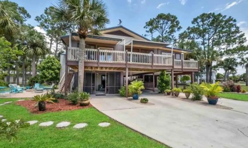 A two-story house surrounded by palm trees and greenery, featuring a spacious driveway and outdoor seating area.