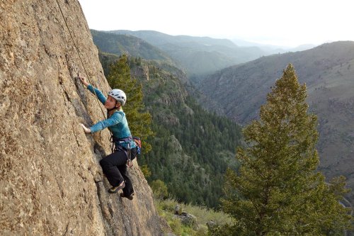 A climber ascends a rocky cliff with mountains and trees in the background, wearing a helmet and climbing gear.