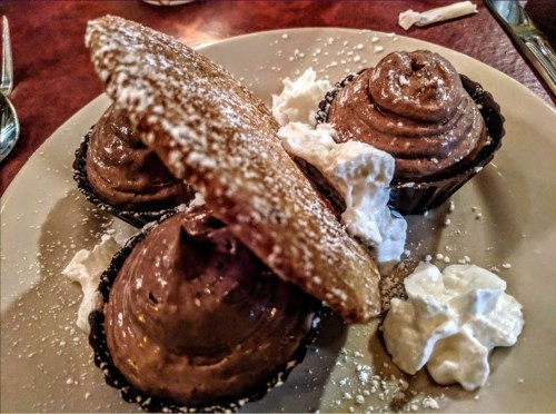 Three chocolate mousse cups topped with whipped cream and a cookie shell, dusted with powdered sugar.