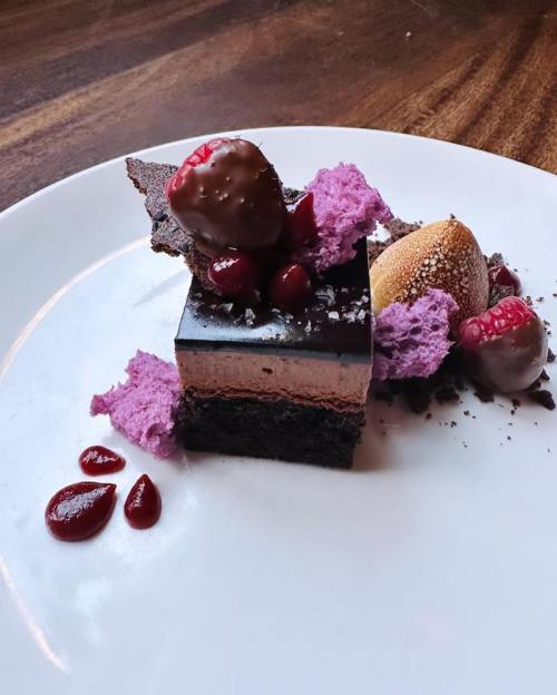 A decadent dessert featuring layers of chocolate, purple mousse, raspberries, and chocolate-covered strawberries on a plate.