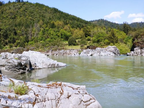 A serene river flows past rocky banks, surrounded by lush green trees and hills under a clear blue sky.