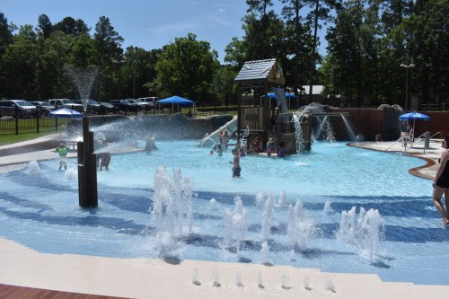 A vibrant water park with children playing in a pool, water slides, and fountains, surrounded by trees and umbrellas.