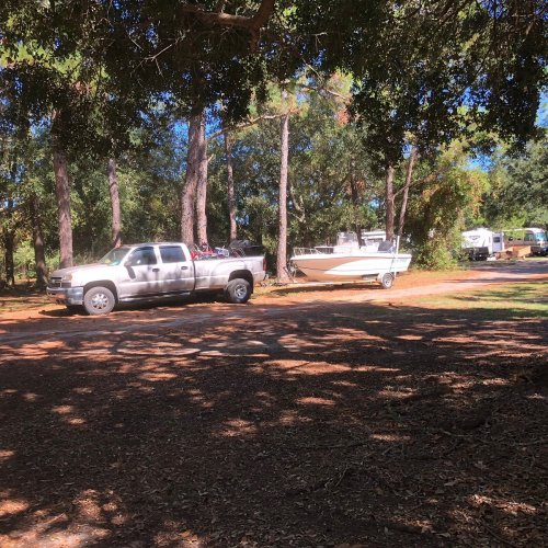 A pickup truck towing a boat parked among trees, with an RV visible in the background on a sunny day.