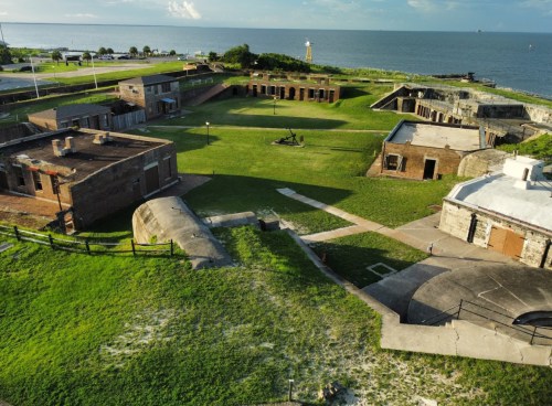 Aerial view of a historic fort with grassy grounds, stone buildings, and a lighthouse in the background by the water.