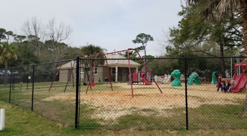 A fenced playground with swings and slides, surrounded by trees and a building in the background.
