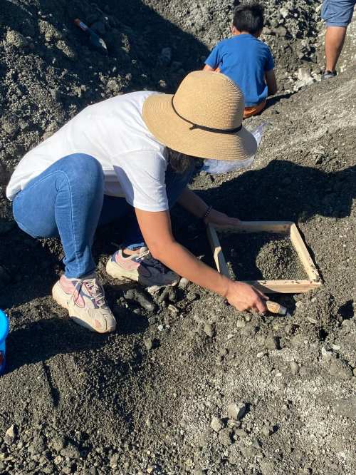 A person in a hat sifts through dirt with a wooden frame, while a child works in the background.