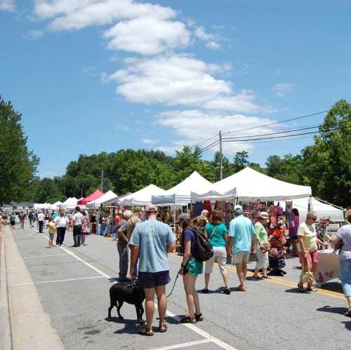 A bustling outdoor market with people browsing stalls under white tents on a sunny day. Trees and blue sky in the background.
