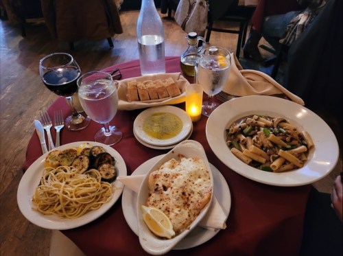 A table setting with pasta, grilled vegetables, a fish dish, bread, and drinks, all arranged on a red tablecloth.