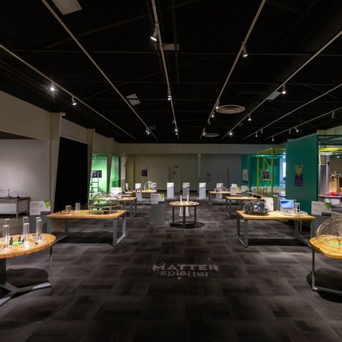 A spacious exhibit room with wooden tables displaying science experiments and informational signs in a dimly lit setting.