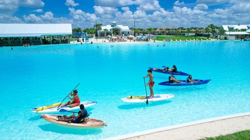People kayaking and paddleboarding in a bright blue lagoon with a sunny sky and palm trees in the background.