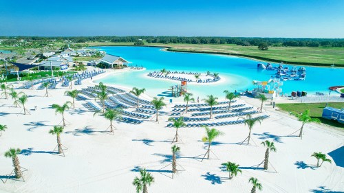 Aerial view of a beach resort with palm trees, lounge chairs, and a turquoise lagoon surrounded by green fields.