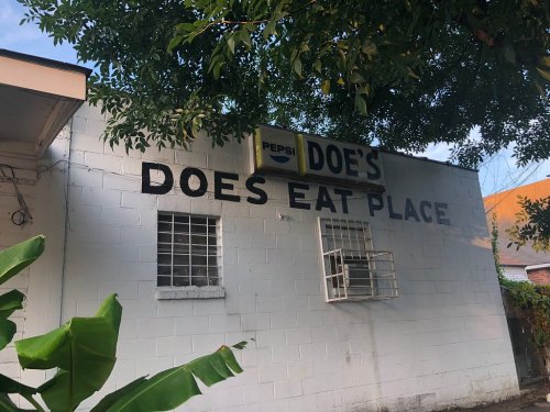 A white wall with the text "Doe's Does Eat Place" and a Pepsi sign, surrounded by greenery.