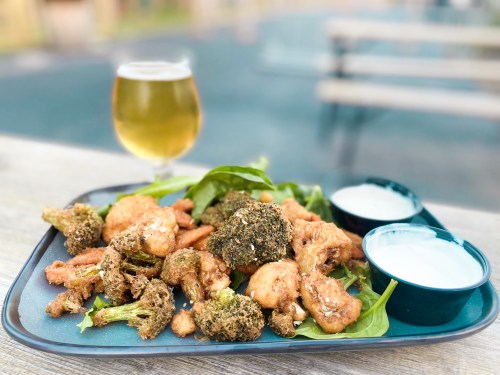 A platter of crispy fried vegetables, including broccoli and cauliflower, served with dipping sauces and a glass of beer.