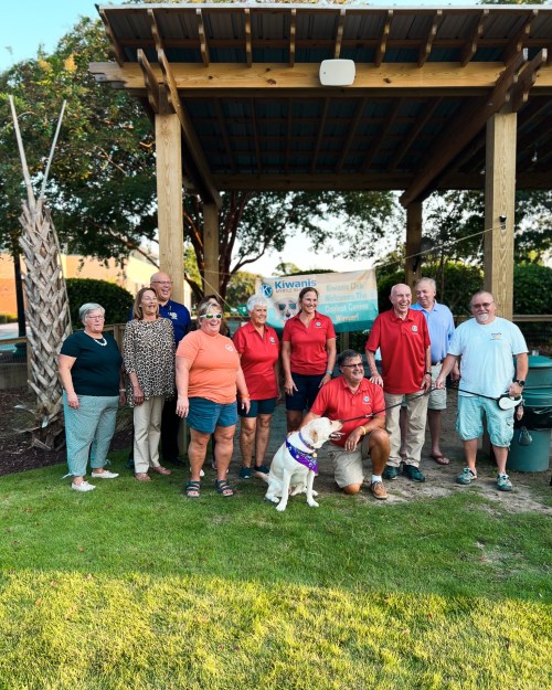 A group of people in red shirts and casual attire pose together outdoors with a dog, under a wooden structure.