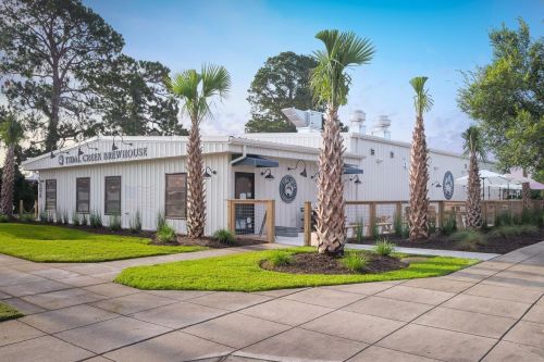 A modern brewery with palm trees, a white exterior, and a landscaped lawn, set against a clear blue sky.