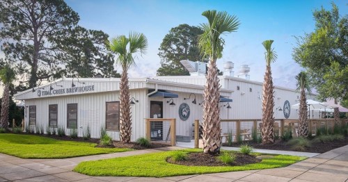 A modern brewery with a white exterior, palm trees, and outdoor seating in a landscaped area.