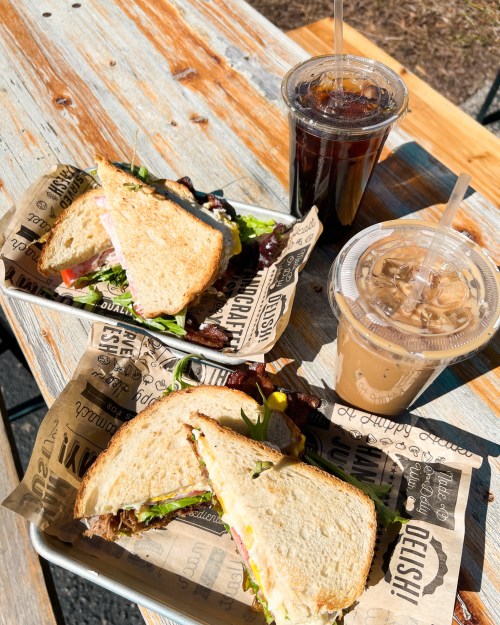 Two sandwiches on paper trays with drinks, set on a rustic wooden table in bright sunlight.