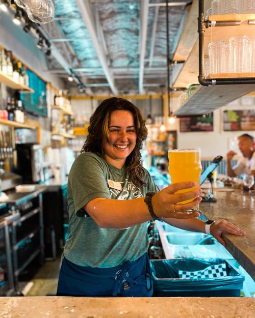 A smiling bartender holds up a glass of beer at a lively bar, showcasing a vibrant atmosphere.