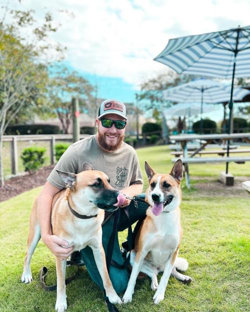 A man in sunglasses sits on grass with two dogs, smiling in a park with picnic tables and umbrellas in the background.