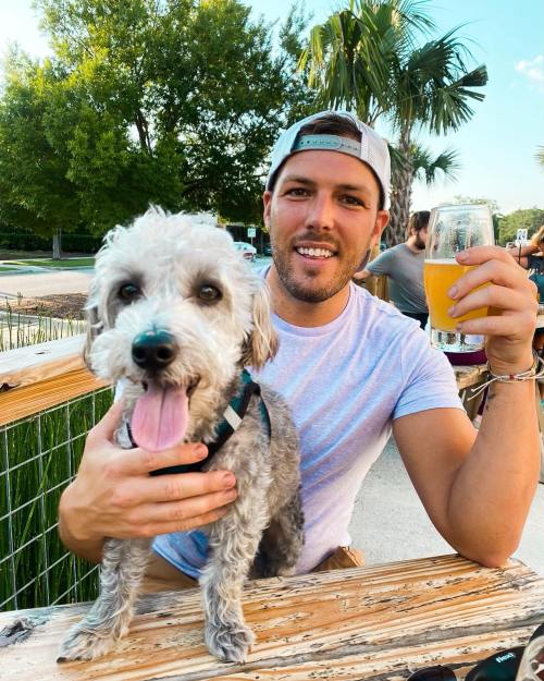 A smiling man holds a drink while sitting at a table with a happy dog beside him, surrounded by greenery.