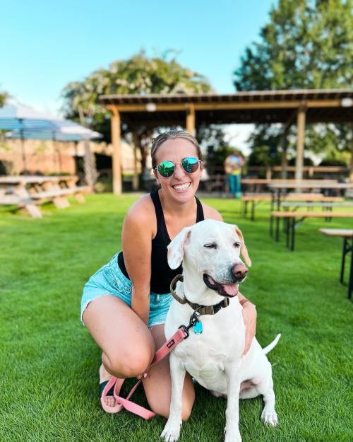 A smiling woman in sunglasses kneels beside a white dog on green grass, with picnic tables in the background.
