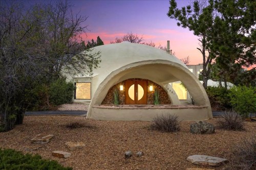 A unique dome-shaped house with a curved entrance, surrounded by desert landscaping and a colorful sunset sky.