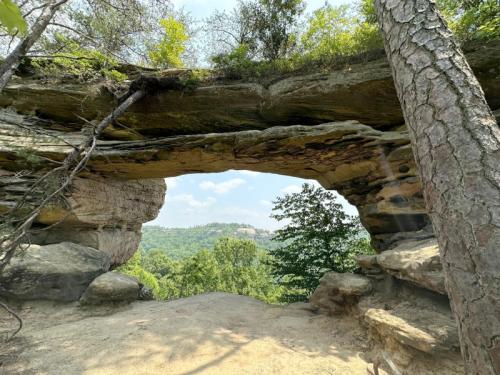 A natural rock arch frames a scenic view of lush green hills and a blue sky in the background.