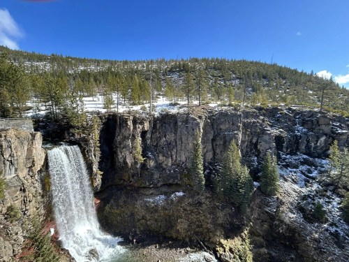 A scenic waterfall cascades down rocky cliffs, surrounded by evergreen trees and a clear blue sky.