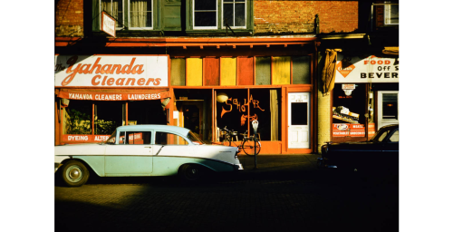 A vintage street scene featuring a light blue car parked outside colorful storefronts, including a cleaners and a food shop.