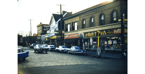 A vintage street scene with classic cars parked outside shops, featuring storefronts and pedestrians on a cobblestone road.