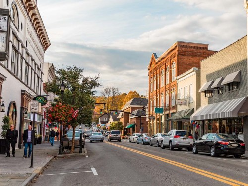 A charming small-town street lined with shops, trees, and parked cars under a cloudy sky.