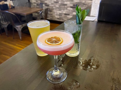 Three drinks on a bar: a pink cocktail with a lemon slice, a clear mint drink, and a pint of beer.