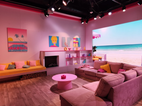 A vibrant living room with pink accents, colorful artwork, and a beach view on a large screen. Cozy seating and decor.