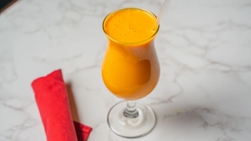 A tall glass of bright orange drink with a straw, placed on a marble surface next to a red napkin.