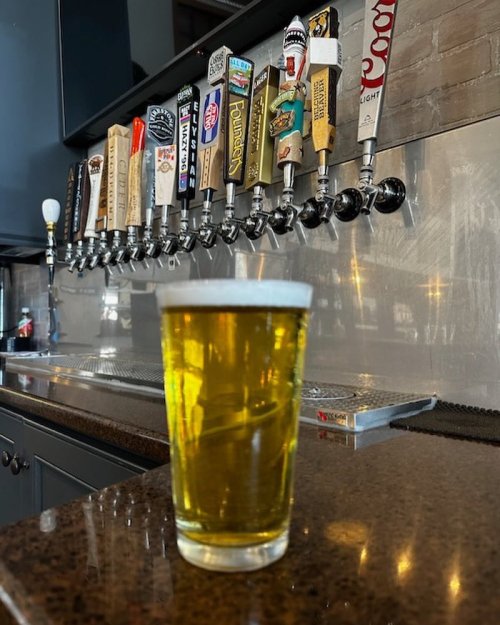 A glass of beer sits on a bar counter in front of a row of colorful beer taps.