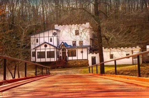 A wooden bridge leads to a castle-like building surrounded by trees in a serene, wooded area.