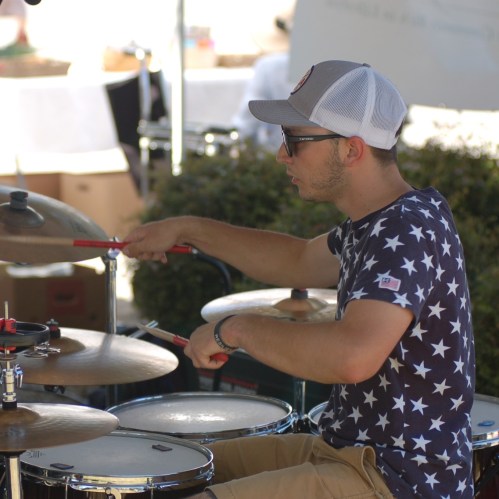 A young man in a star-patterned shirt plays drums outdoors, focused on his performance.