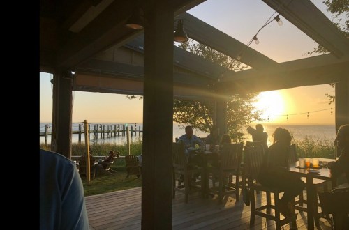 A sunset view from a cozy outdoor restaurant, with people dining and a pier visible in the background.