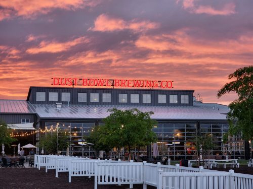 A brewery building with "Dust Bowl Brewing Co" sign against a colorful sunset sky, surrounded by greenery and outdoor seating.
