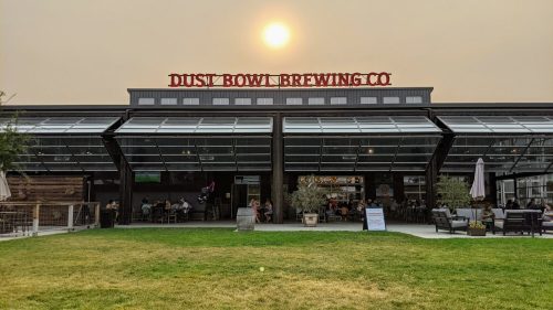 Dust Bowl Brewing Co. building with a large sun setting behind it, outdoor seating, and people enjoying the space.