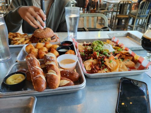 A table with a burger, fries, pretzels, nachos, and drinks in a casual dining setting.