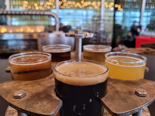 A flight of six craft beers in clear glasses, displayed on a metal holder, with a blurred bar background.