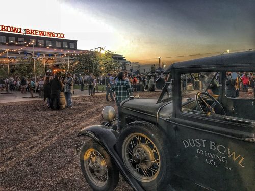 A vintage car in the foreground with a brewery and crowd enjoying an outdoor event at sunset in the background.